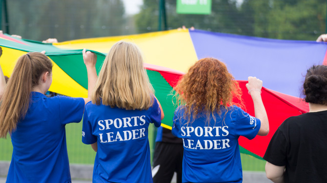 Image shows a group of pupils in Sports Leader tops doing an exercise with a parachute
