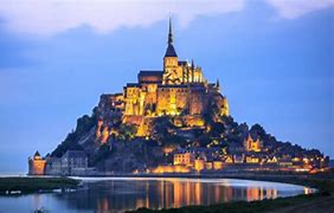 Image shows a photo of Mont Saint Michel illuminated at dusk, and reflected in the sea